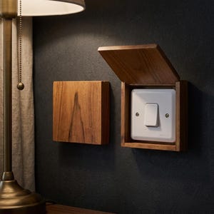 Op de afbeelding: Twee vierkante houten afdekplaatjes gemonteerd op een donkere muur. Eén plaatje is gesloten, terwijl de andere open staat en een witte rechthoekige lichtschakelaar met een tuimelschakelaar en twee zilveren schroeven onthult. Zichtbare houtnerf.