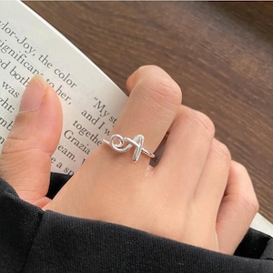 Könnte beinhalten: Ein silberfarbener Ring mit einem stilisierten, kursiven Buchstaben A als zentrales Designelement. Das Band ist dünn und glatt, wobei der Buchstabe A aus einem durchgehenden, abgerundeten Metalldraht geformt ist, der auf dem Finger sitzt.