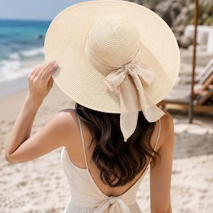 May include: A wide-brimmed beige straw hat featuring a matching fabric ribbon tied in a bow at the back. The hat is paired with a sleeveless, backless beige dress that also features a fabric tie closure at the center of the back.
