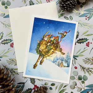 May include: A Christmas card featuring a whimsical illustration of Santa Claus and elves riding a goat through a snowy landscape. The card has a white envelope and is set against a festive background with pine cones and greenery.