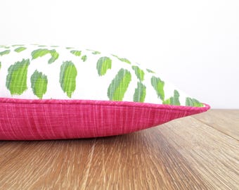 May include: Decorative pillow with a white background and green leaf-like pattern. The bottom of the pillow is a solid magenta color. The pillow is on a wooden surface.