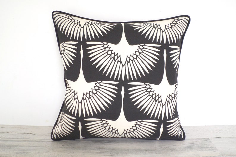 May include: Square decorative pillow with a black background and a repeating white crane design. The pillow has a black border and is placed against a white background.