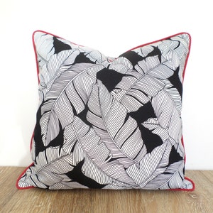May include: Square decorative pillow with a black background and a white leaf pattern. The pillow has a red trim around the edges. The pillow is made of fabric and is designed for home decor.