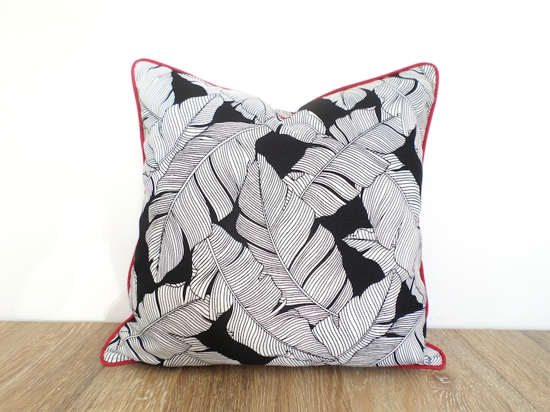 May include: Square decorative pillow with a black background and a white leaf pattern. The pillow has a red trim around the edges. The pillow is made of fabric and is designed for home decor.