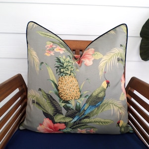 May include: Gray decorative pillow with a tropical design featuring a pineapple, parrot, and floral elements. The pillow has a dark blue trim and is placed on a wooden chair with a navy blue cushion.