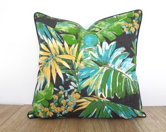 May include: Square decorative pillow with a tropical leaf pattern in shades of green, teal, and yellow on a dark gray background. The pillow has a navy blue trim and is placed on a wooden surface.
