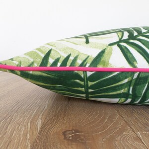 Op de afbeelding: Een decoratief kussen met een tropisch bladerpatroon in groen en wit. Het heeft een helder roze bies. Het kussen ligt op een houten oppervlak, wat een contrast cre&euml;ert.