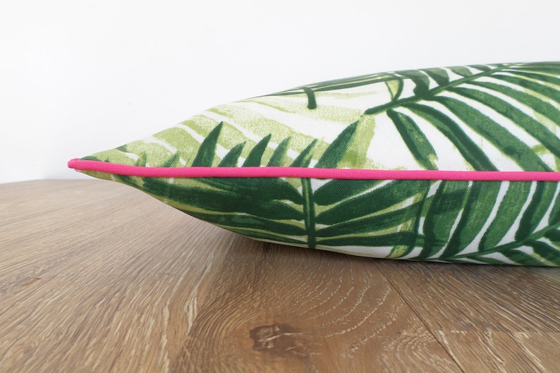 Op de afbeelding: Een decoratief kussen met een tropisch bladerpatroon in groen en wit. Het heeft een helder roze bies. Het kussen ligt op een houten oppervlak, wat een contrast cre&euml;ert.