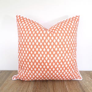 May include: A square throw pillow featuring a vibrant orange background with a repeating white leaf-like pattern. The pillow is finished with white piping along the edges, creating a clean and defined border for the overall square shape.