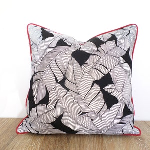 May include: Square throw pillow with a black background and a white leaf pattern. The pillow has a red border. The pillow is on a wooden surface.
