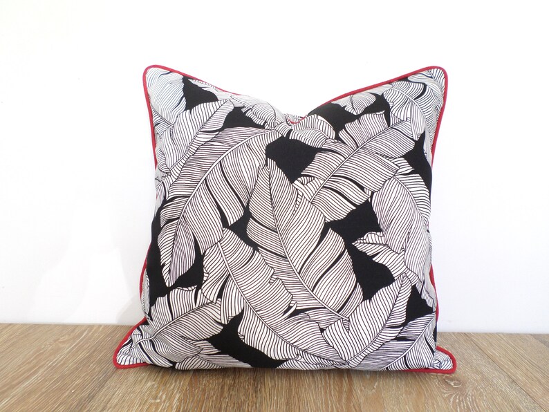 May include: Square throw pillow with a black background and a white leaf pattern. The pillow has a red border. The pillow is on a wooden surface.