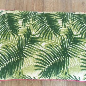 Op de afbeelding: Rechthoekig decoratief kussen met een tropisch palmbladpatroon in groen- en wittinten. Het kussen heeft een helder roze rand, wat een vleugje kleur toevoegt. Geplaatst op een houten oppervlak.