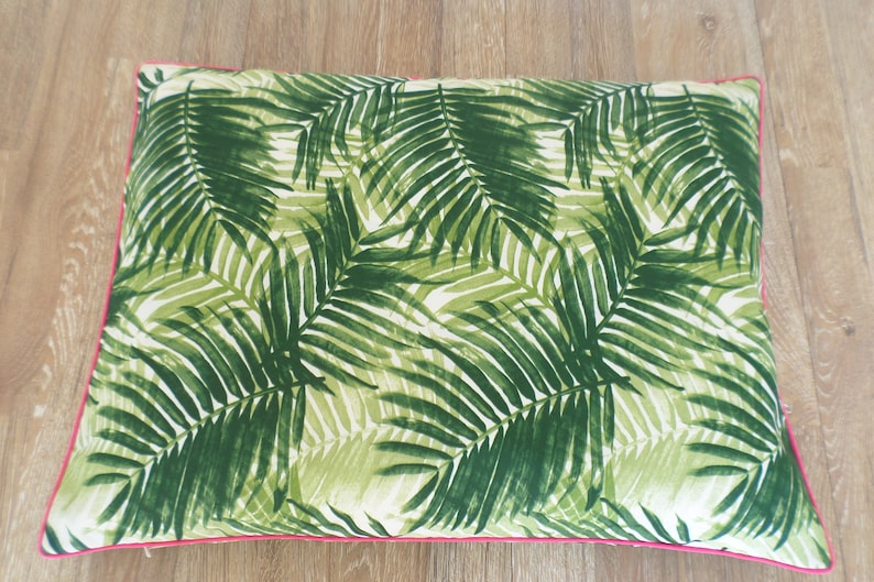 Op de afbeelding: Rechthoekig decoratief kussen met een tropisch palmbladpatroon in groen- en wittinten. Het kussen heeft een helder roze rand, wat een vleugje kleur toevoegt. Geplaatst op een houten oppervlak.