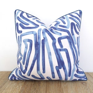 May include: Square decorative pillow with a white background and a blue abstract design. The pillow has a dark blue trim. The pattern consists of blue lines of varying widths and shades, creating a modern look. Placed on a wooden surface.