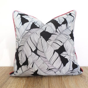 May include: Square throw pillow with a black background and a white leaf pattern. The pillow has a red trim. The pillow is placed on a wooden surface against a white background. Home decor item.