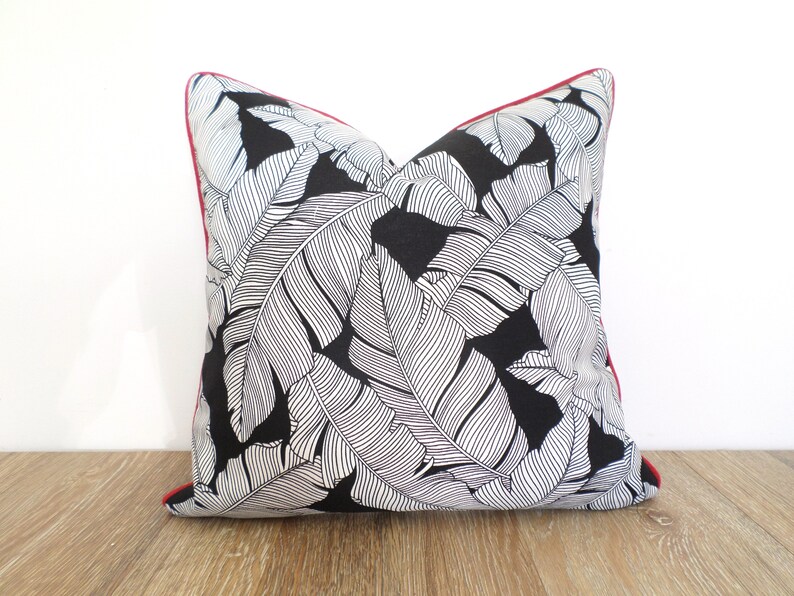 May include: Square throw pillow with a black background and a white leaf pattern. The pillow has a red trim. The pillow is placed on a wooden surface against a white background. Home decor item.