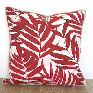 May include: Square decorative pillow with a red leaf pattern on a white background. The pillow has a beige border. The pillow is placed on a wooden surface.