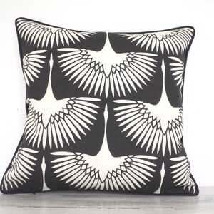 May include: Square decorative pillow with a black background and a repeating pattern of white crane illustrations. The pillow has a black border and is likely made of fabric. The design is modern and suitable for home decor.