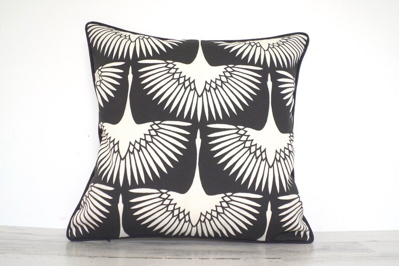 May include: Square decorative pillow with a black background and a repeating pattern of white crane illustrations. The pillow has a black border and is likely made of fabric. The design is modern and suitable for home decor.