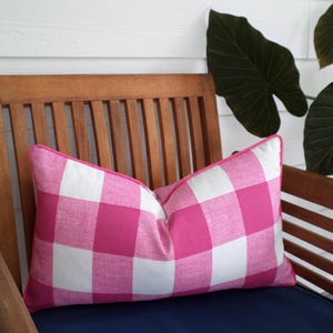 May include: A pink and white checkered pillow with a pink border rests on a wooden chair with a navy blue cushion. The pillow is rectangular and has a classic gingham pattern. The chair is made of wood and has a slatted back.