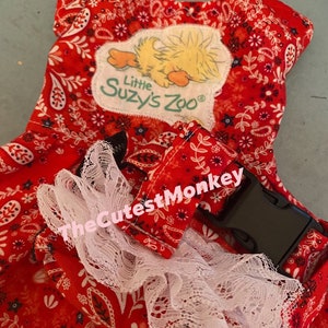 May include: Red and white paisley baby carrier with a white lace trim and a black buckle. The carrier has a patch with a yellow chick and the text "Little Suzy's Zoo".