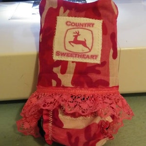 May include: Pink and white camouflage fabric doll dress with a lace trim and a white patch that says "COUNTRY SWEETHEART" with a red deer silhouette.