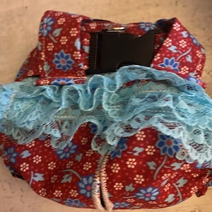 May include: A red fabric baby carrier with a blue floral pattern and a black buckle. The carrier has a blue lace trim.