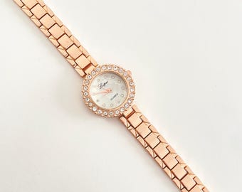 May include: Rose gold wristwatch with a round face and a bezel encrusted with small, clear crystals. The watch has a white dial with delicate hands and the word "Quartz" printed on it. The band is made of rose gold links.
