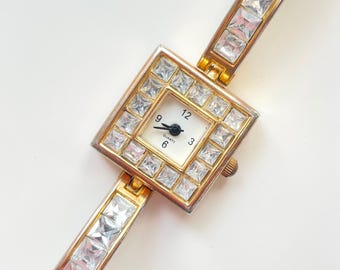 Può includere: Un orologio al quarzo color oro con quadrante quadrato e cinturino impreziosito da cristalli trasparenti a taglio quadrato. Il quadrante dell'orologio è bianco con lancette e numeri neri. L'orologio è in stile vintage.