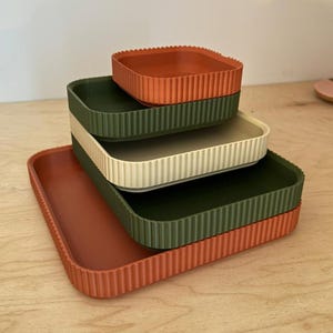 May include: Four rectangular trays in terracotta, olive green, and cream are stacked. The trays have a ribbed design around the edges. The largest tray is at the bottom. The trays are arranged on a wooden surface.