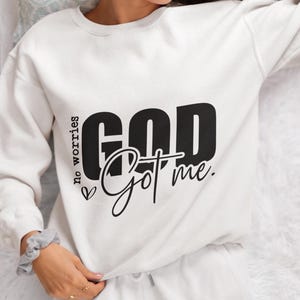 May include: White sweatshirt featuring the text "GOD Got me" in black, with "no worries" written vertically on the left. The sweatshirt has a crew neck and long sleeves. The person is wearing white sweatpants.