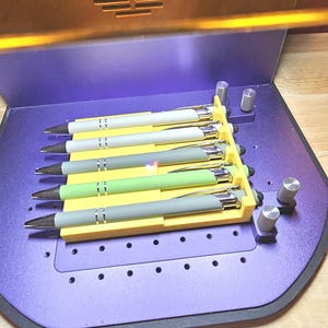 May include: A collection of six pens in a yellow holder, each with a different colored barrel: white, gray, green, and gray. The pens have silver accents and black tips. The holder sits on a purple surface with silver knobs.