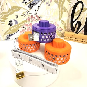 May include: Three colourful tape measure holders in orange and purple, with a honeycomb design. A white tape measure extends from one holder, showing cm measurements up to 23 cm. The holders sit on a white surface.