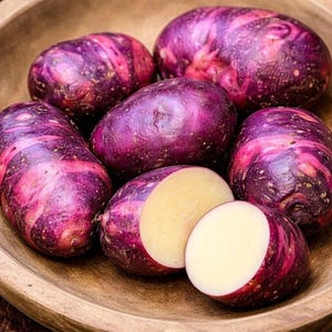 May include: A collection of oval-shaped potatoes with deep purple and magenta mottled skin. One potato is sliced in half, revealing a pale cream-colored interior. The potatoes have a smooth, firm texture and are arranged in a wooden bowl.