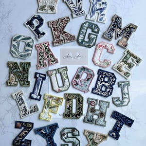 May include: Embroidered fabric letters in a variety of colours and floral designs. Each letter has a white border and is arranged on a white surface. The letters are from A to Z, with a few missing. A small card with text is also visible.