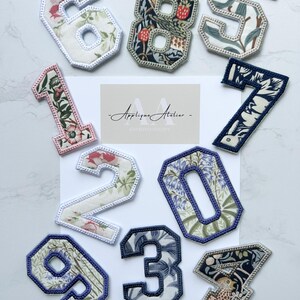 May include: Embroidered fabric numbers with floral patterns, ranging from 0 to 9. Each number has a contrasting border and decorative stitching. The image also features the text "- Applique Atelier - EMBROIDERY" and a website address, all in a decorative font.