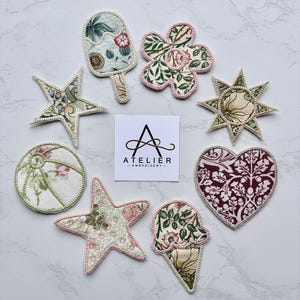 May include: A collection of eight fabric patches featuring various shapes including stars, a heart, a flower, a beach ball, and ice cream treats, all with floral patterns and stitched edges. A square card in the centre displays the text ATELIER EMBROIDERY.