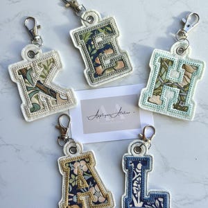 May include: Five embroidered initial keyrings with floral patterns. The letters K, E, H, A, and L, are displayed on white backing with silver clasps. The keyrings feature blue, green, and peach floral designs. The keyrings are ideal for personalising bags or keys.