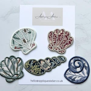 May include: Five embroidered seashell patches in various colours and patterns are displayed on a white background. The patches include floral and botanical designs in shades of blue, red, and green. The text "Applique Atelier" and a website address are also visible.