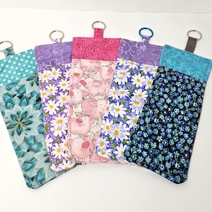 May include: Five rectangular fabric pouches with metal key rings attached to the top. The pouches feature various patterns including teal mandalas, purple daisies, pink pigs, white daisies on purple, and small blue flowers on a black background.