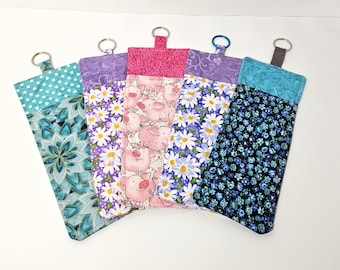 May include: Five rectangular fabric pouches with metal key rings attached to the top. The pouches feature various patterns including teal mandalas, purple daisies, pink pigs, white daisies on purple, and small blue flowers on a black background.