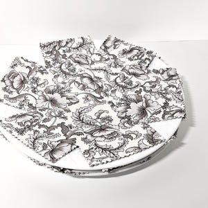 May include: A stack of square fabric napkins featuring a black and white floral pattern with intricate botanical details. The napkins are arranged in a circular formation on top of each other, showcasing the repeating print of large blossoms and leaves.