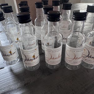 May include: A collection of small, clear glass bottles with black screw-top lids. Each bottle features a white rectangular label with gold accents and the printed text "Five Decades of Jánika Fabulous" in a decorative script font.