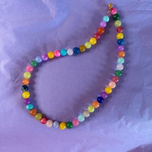 Könnte beinhalten: Eine bunte Perlenkette mit einer Vielzahl von Farben, darunter Rot, Orange, Gelb, Grün, Blau, Lila und Pink. Die Perlen sind auf einer goldenen Kette aufgefädelt.