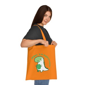 Può includere: Borsa di tela arancione con un'illustrazione di dinosauro verde e il testo "chaos is a superpower".