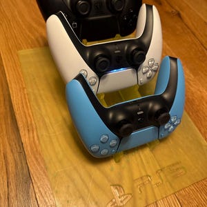 May include: Three PlayStation 5 DualSense controllers, one black, one white, and one light blue, are stacked on a clear plastic stand with the PlayStation logo.