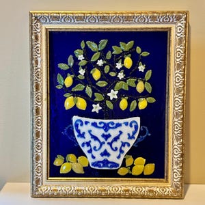 May include: A framed wall art piece featuring a three-dimensional glass lemon tree in a white and blue patterned teacup, set against a deep blue background. The tree has green leaves, white flowers, and yellow lemons, with loose lemons at the base.