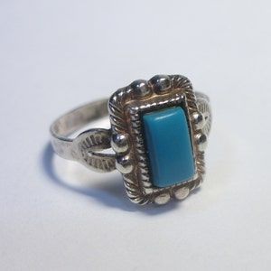 May include: A silver ring with a turquoise stone set in a square bezel. The ring has a decorative band with a southwestern design.