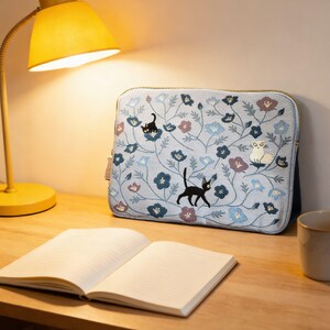Pu&ograve; includere: Una custodia per laptop azzurra con un motivo floreale e gatti neri e bianchi ricamati. La custodia ha una cerniera dorata ed &egrave; appoggiata su una scrivania di legno accanto a un quaderno aperto e una lampada da scrivania gialla.