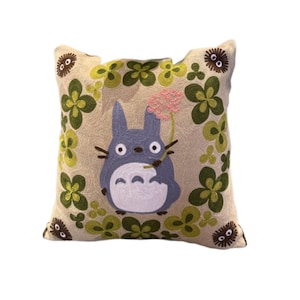 Puede incluir: Cojín cuadrado bordado con un personaje Totoro azul sosteniendo una flor rosa. El cojín tiene un fondo beige con diseños de tréboles verdes y amarillos y pequeños personajes redondos marrones. Un acento decorativo para una cama o sofá.